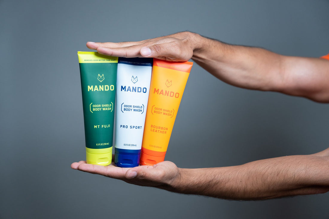 person holding three tubes of mando deodorant upright in between palms against a grey background
