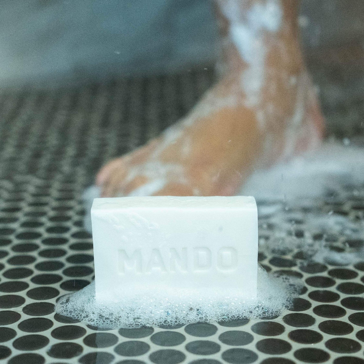 White mando cleansing bar placed on shower floor with persons leg in background 