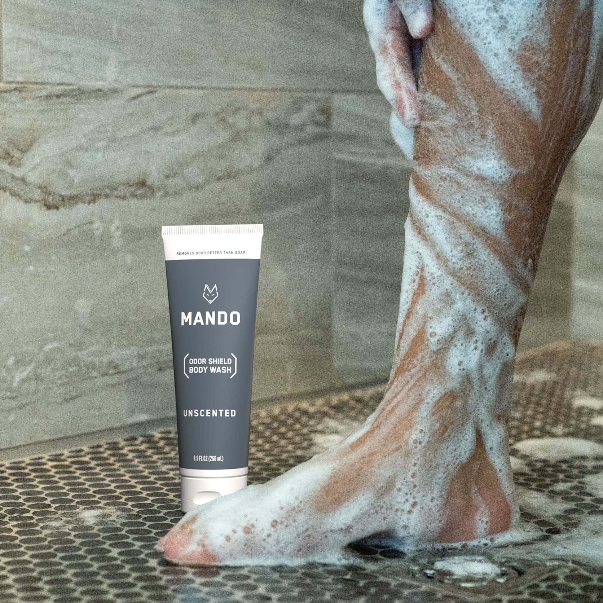 person lathering soap on leg with grey tube of Mando unscented body wash on shower floor