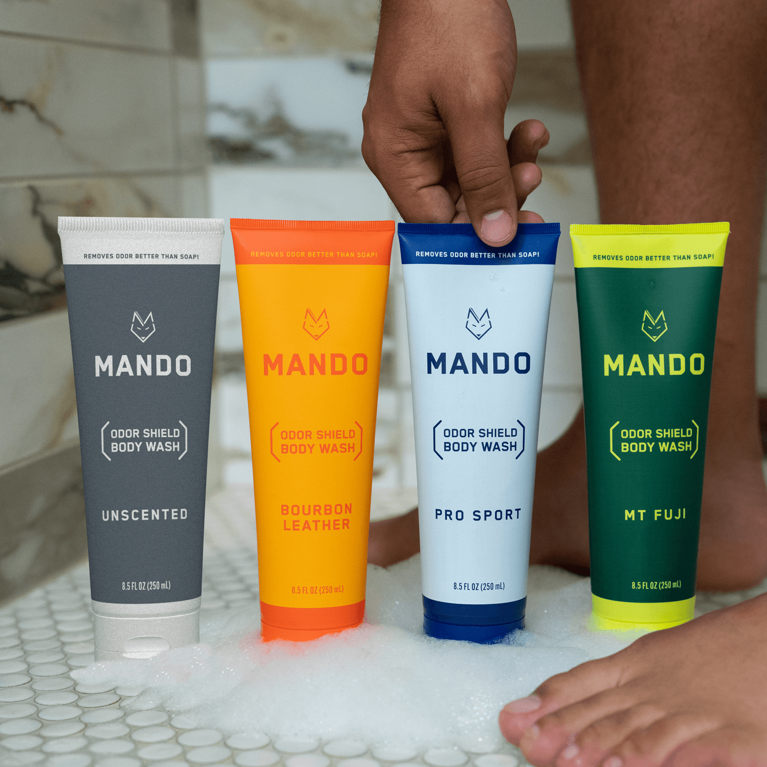 line up of Mando body wash in unscented, bourbon leather, pro sport and mt fuji placed on shower floor with person in background