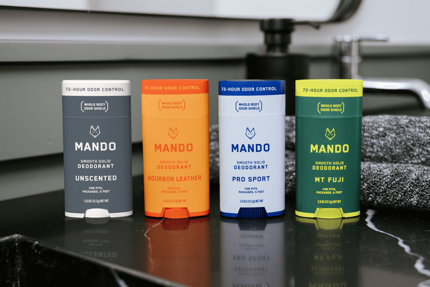 line up of 4 Mando solid deodorants in unscented, bourbon leather, pro sport and mt fuji placed on a counter top with a grey towel in the background